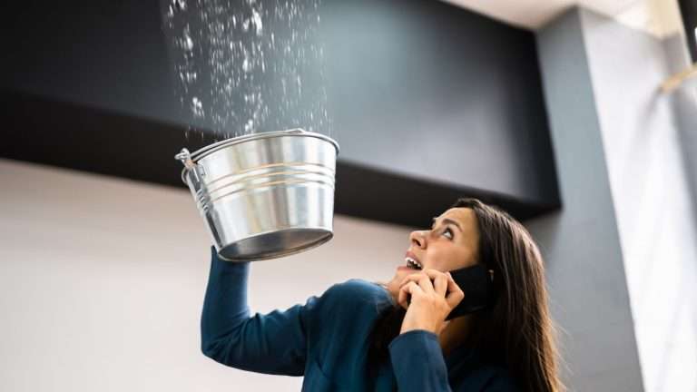 Worried woman catches ceiling leak with bucket while calling for water damage cleanup.